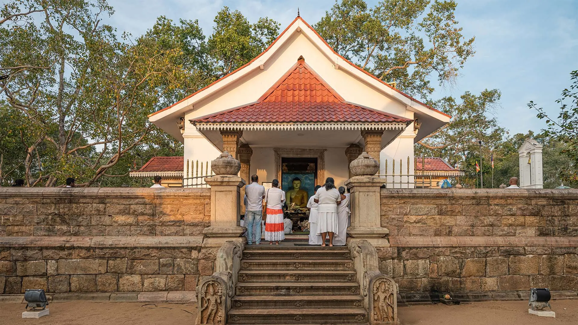 Jaya Sri Maha Bodhi temple with visitors at the entrance amidst lush trees.