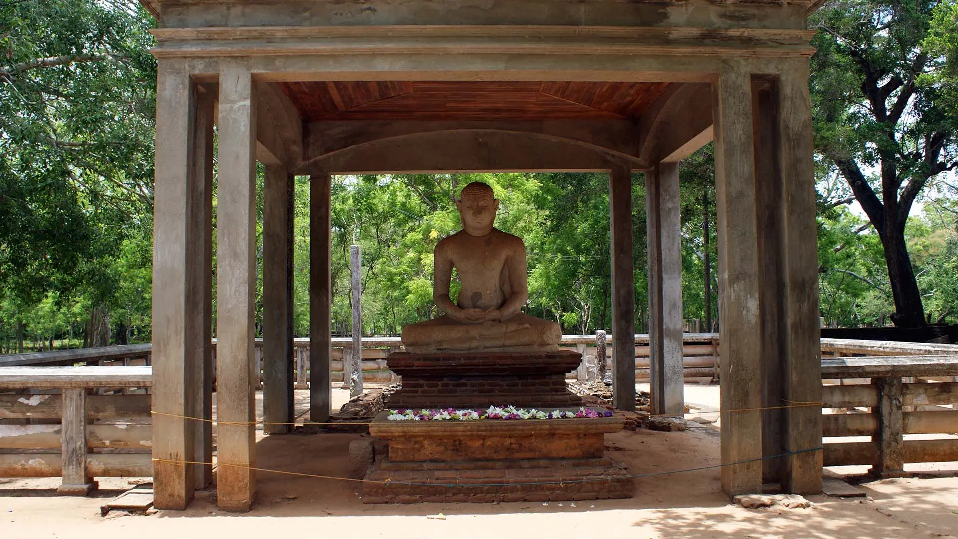 Samadhi Buddha Statue in a stone pavilion adorned with flowers amidst lush greenery.