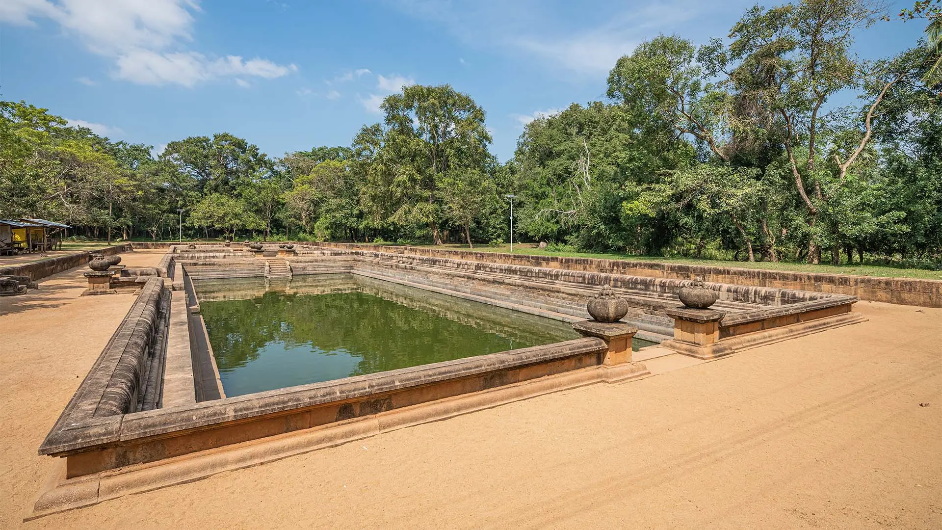 Ancient Kuttam Pokuna with stone walls amidst lush greenery under a clear blue sky.