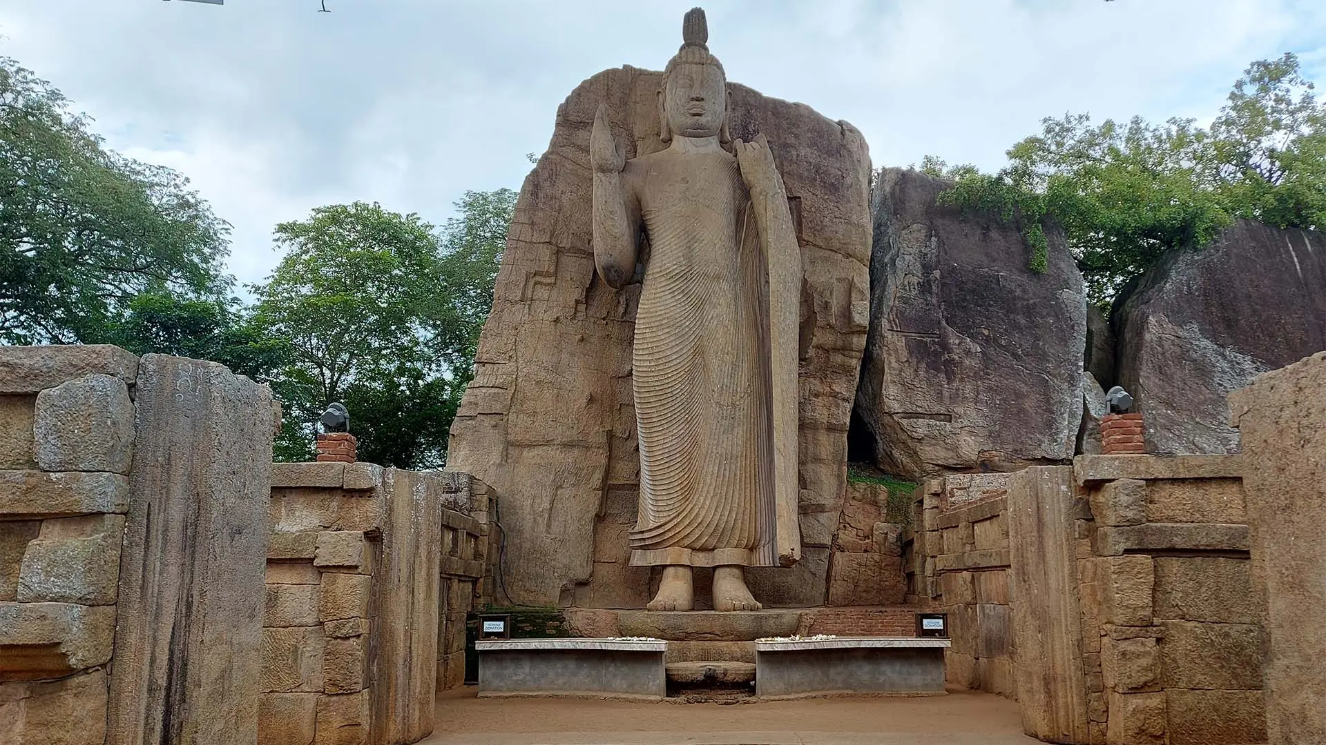 Towering Avukana Buddha Statue carved in rock with raised hand amidst ancient stone structures and lush greenery.