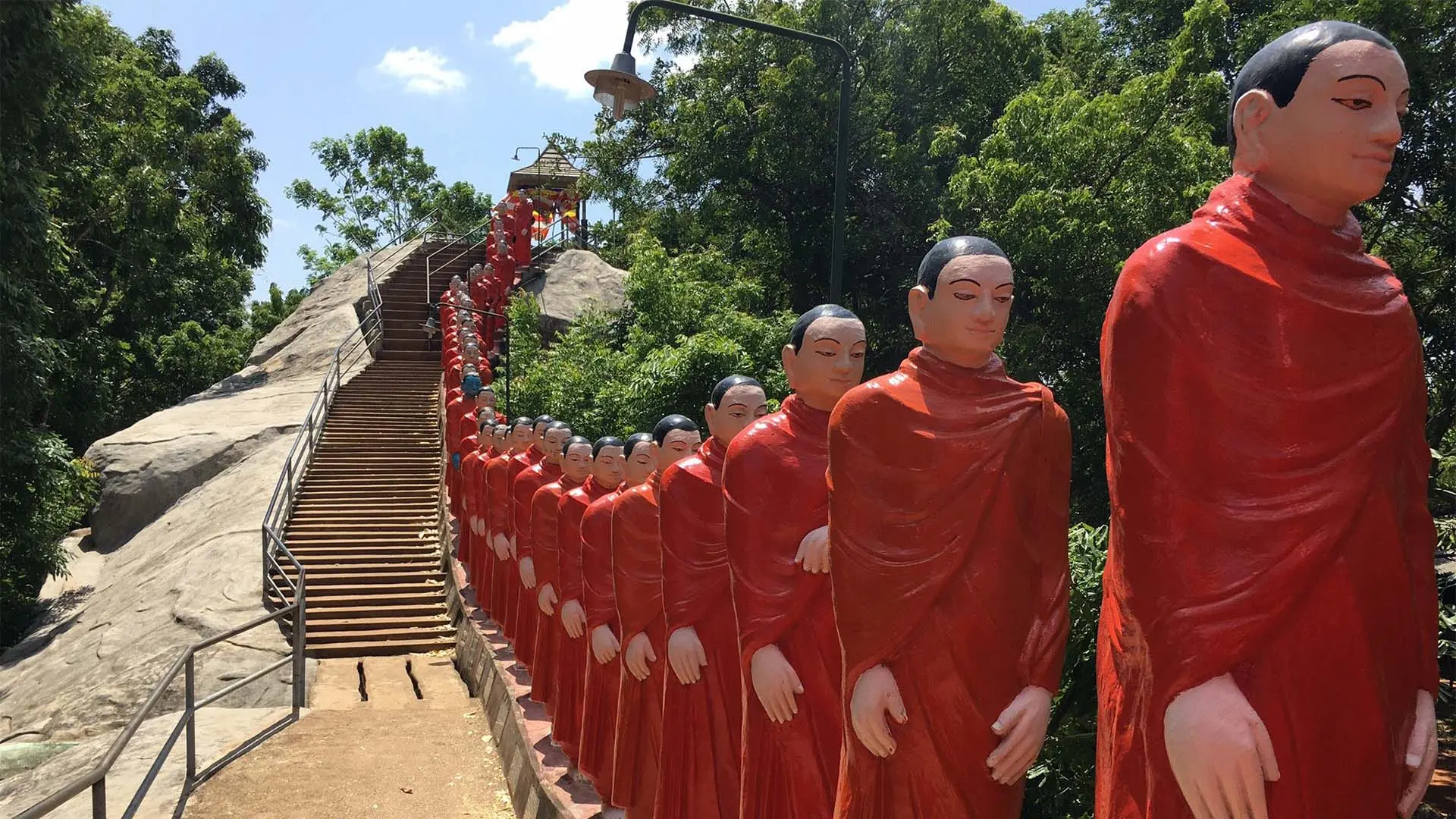 Nellikulama Temple with a staircase lined by red-robed Buddha statues amidst lush greenery under a clear sky.
