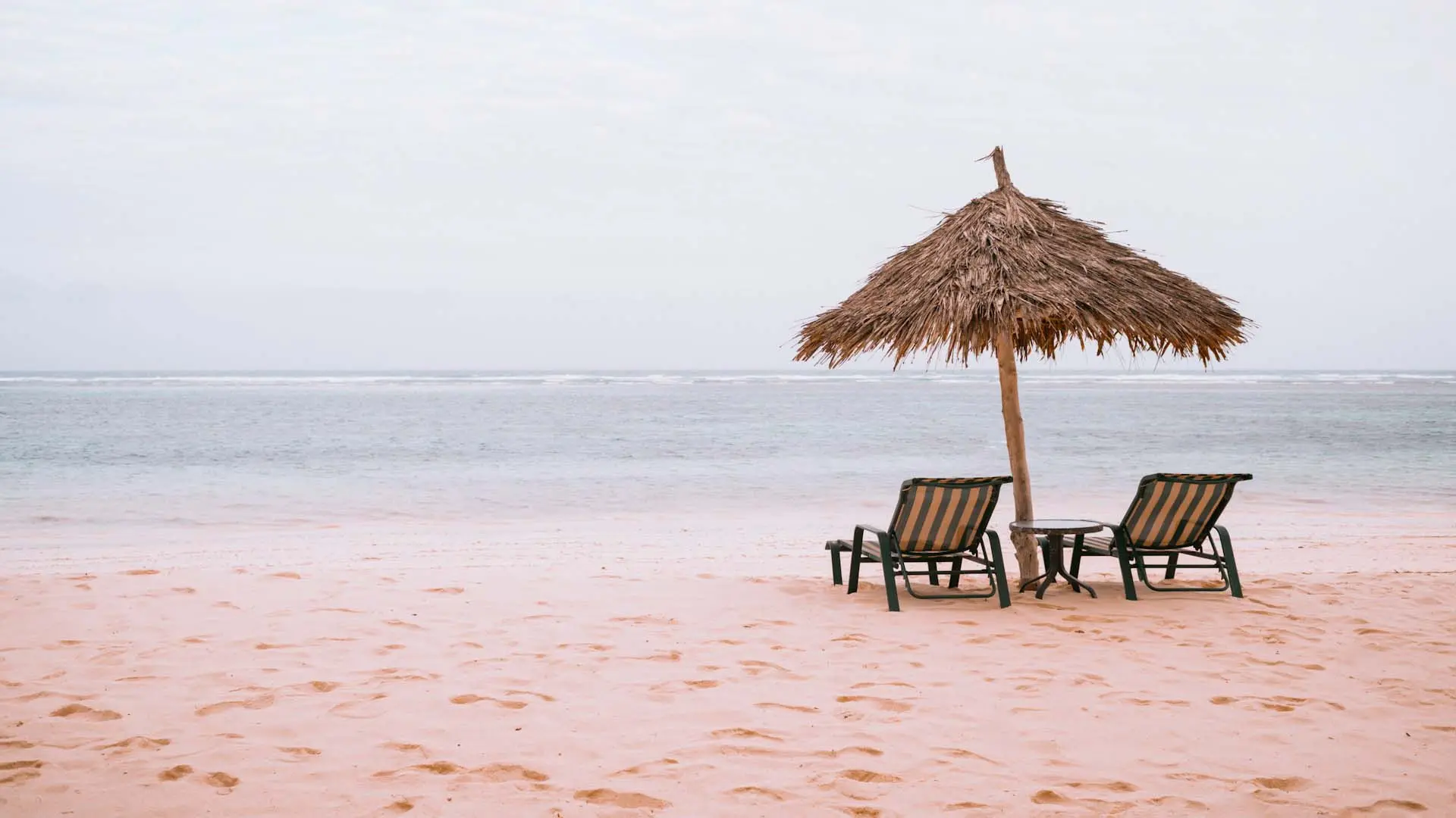 Bentota Beach with tranquil sandy shore, lounge chairs under a thatched umbrella, and calm ocean waters under a cloudy sky.