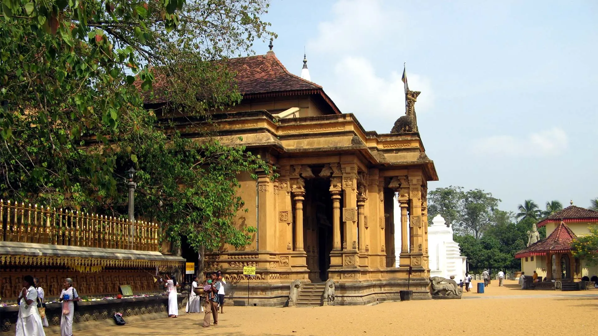 Kelaniya Raja Maha Viharaya with intricate stone architecture and red-tiled roof, surrounded by visitors under a partly cloudy sky.