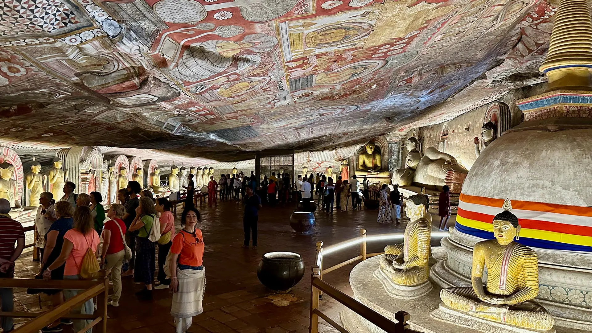 Inside Dambulla Royal Cave Temple with intricate ceiling paintings and Buddha statues among visitors.