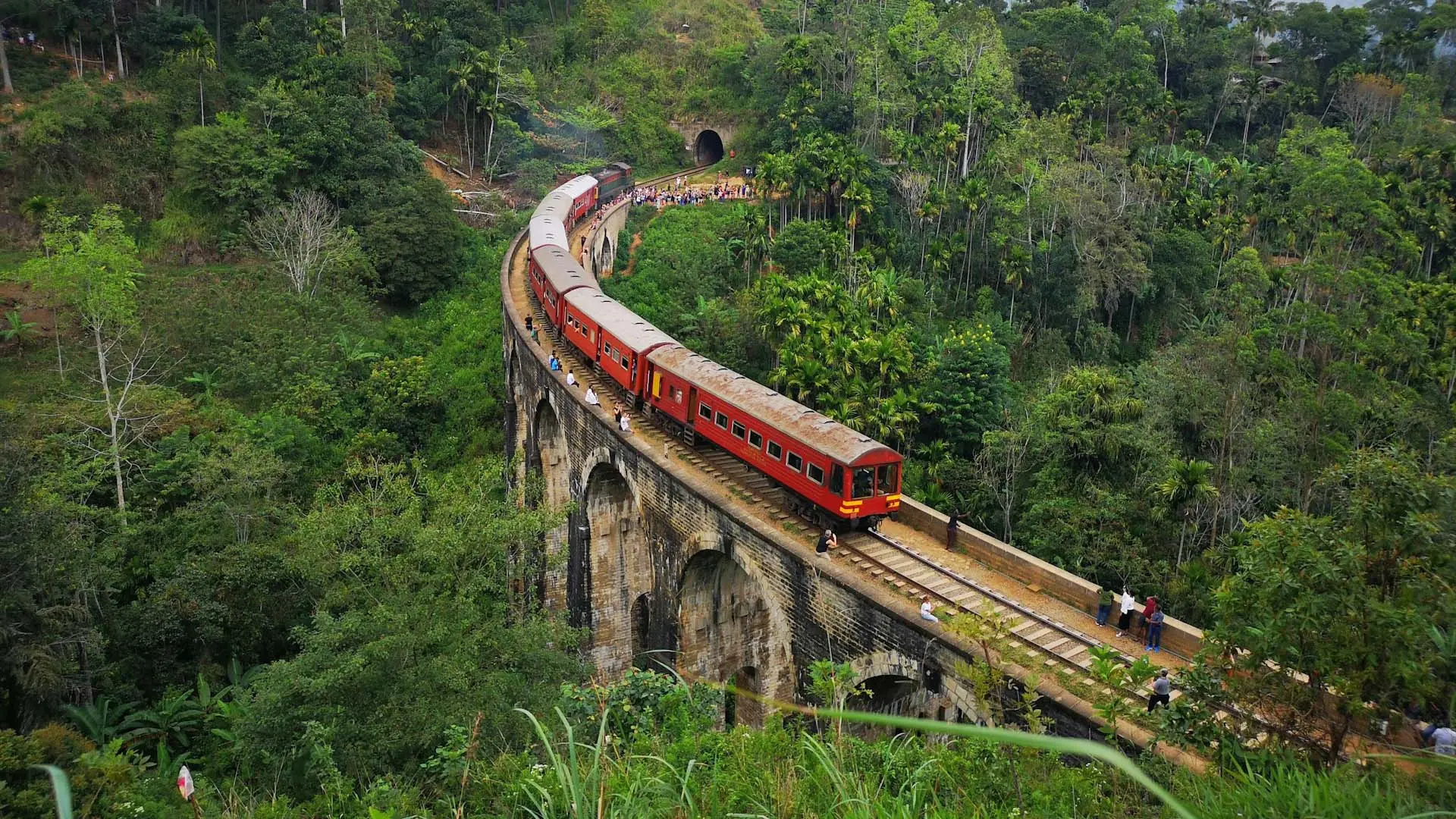 Red train crossing the historic Nine Arch Bridge amidst lush green hills.