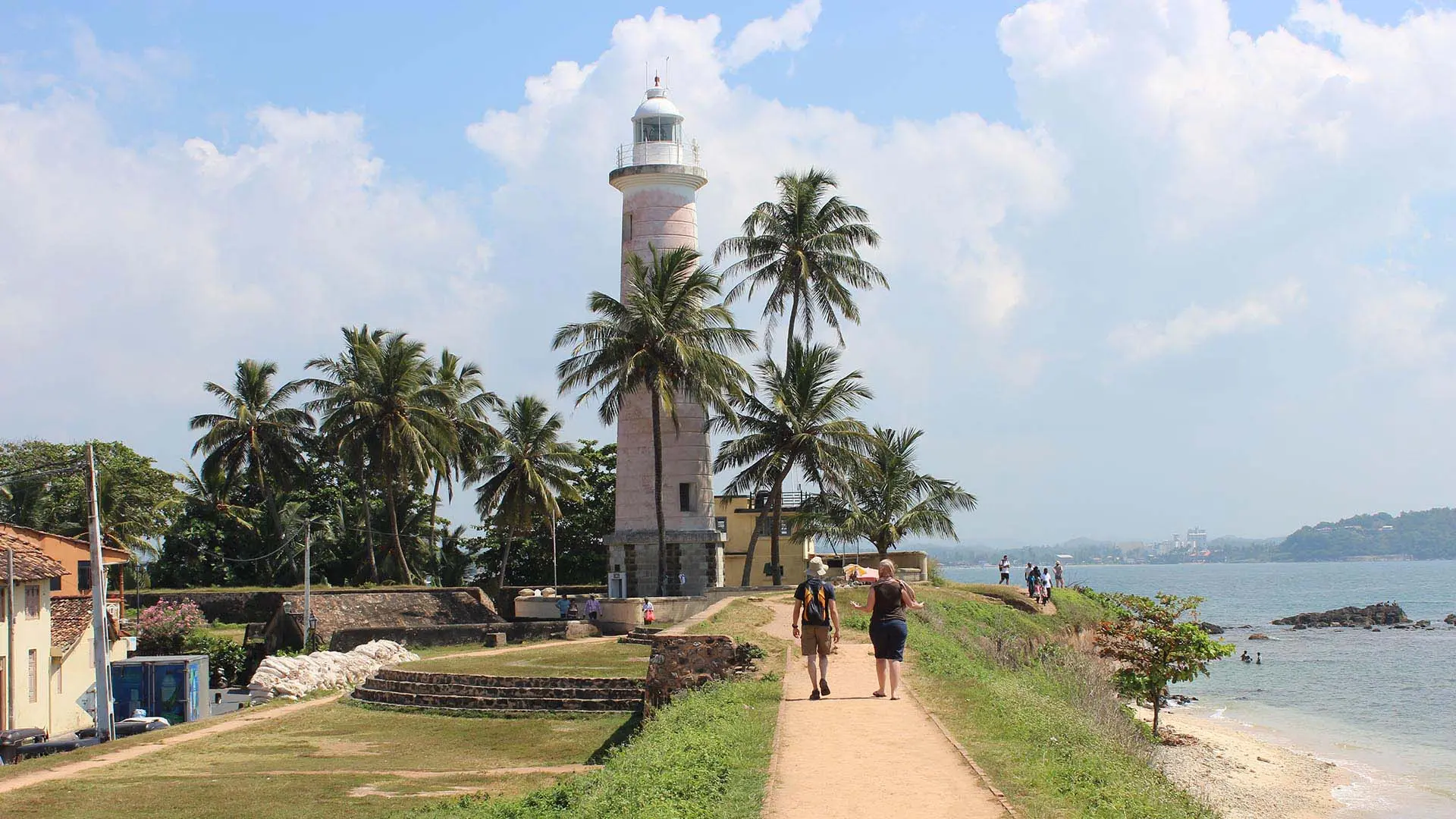 Galle Dutch Fort with historic lighthouse, palm trees, and a coastal pathway with visitors under a partly cloudy sky.