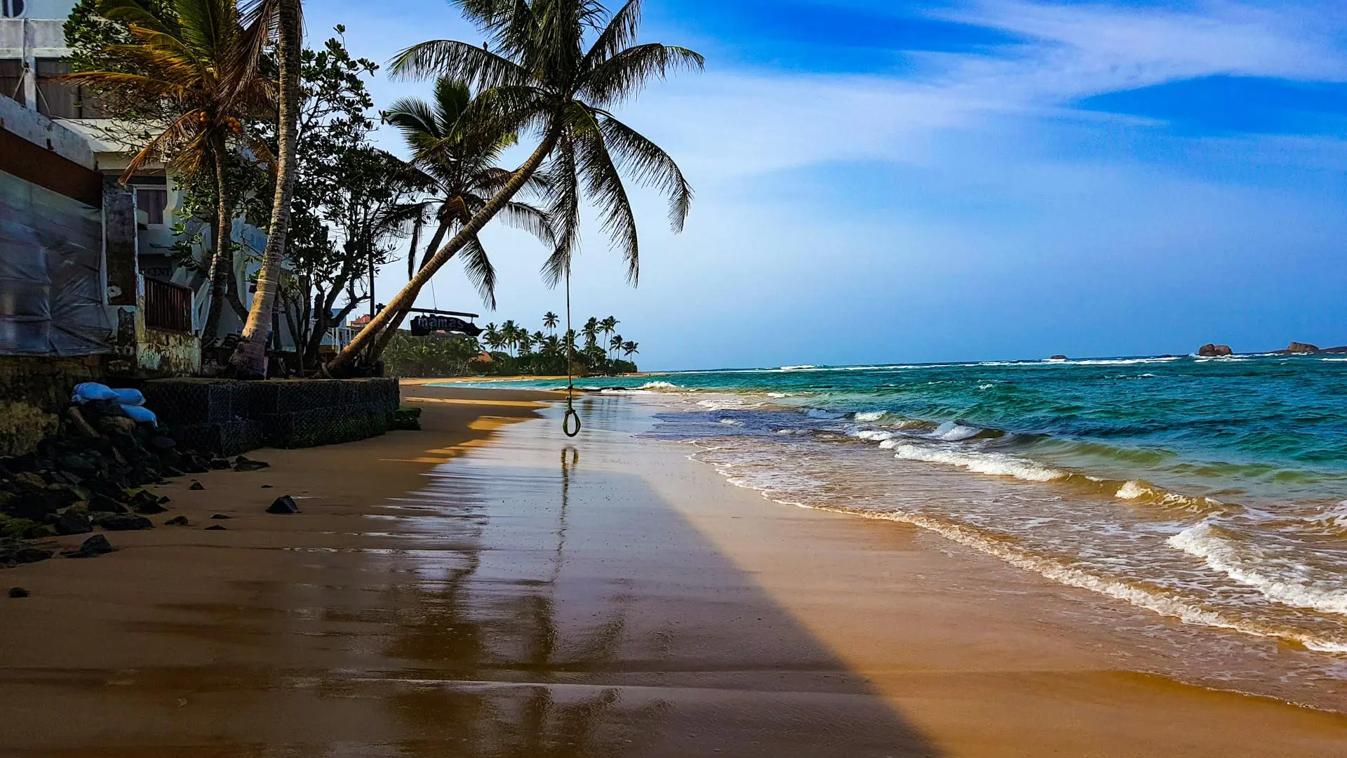 Hikkaduwa Beach with serene sandy shoreline, palm trees, turquoise waters, and distant rock formations under a clear blue sky.