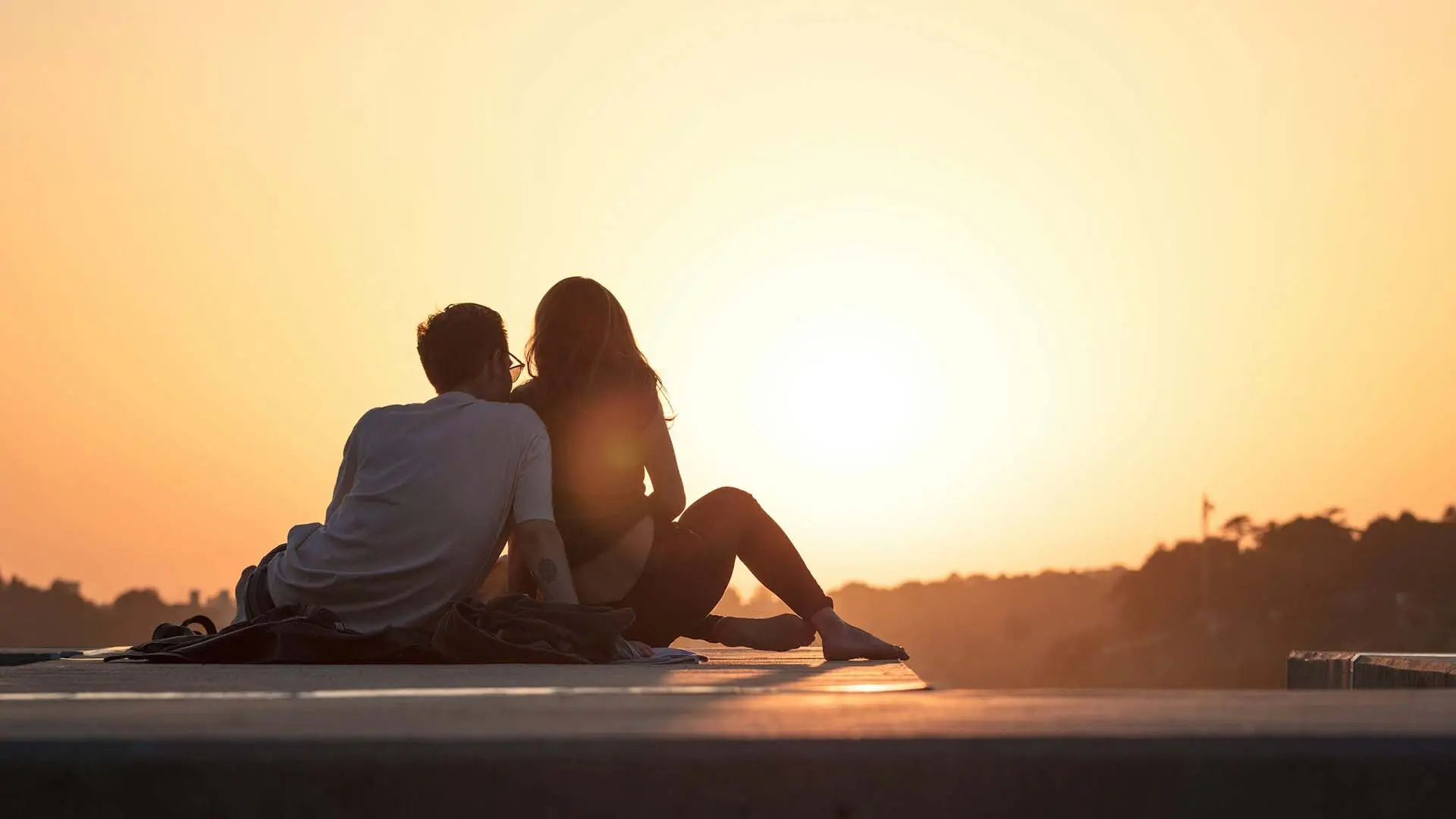 A couple silhouetted against a vibrant sunset and distant greenery.