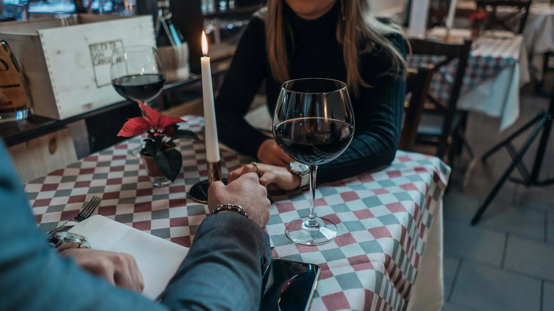 Candlelight dinner with a couple holding hands, lit candle, red wine, and checkered tablecloth in a cozy restaurant setting.