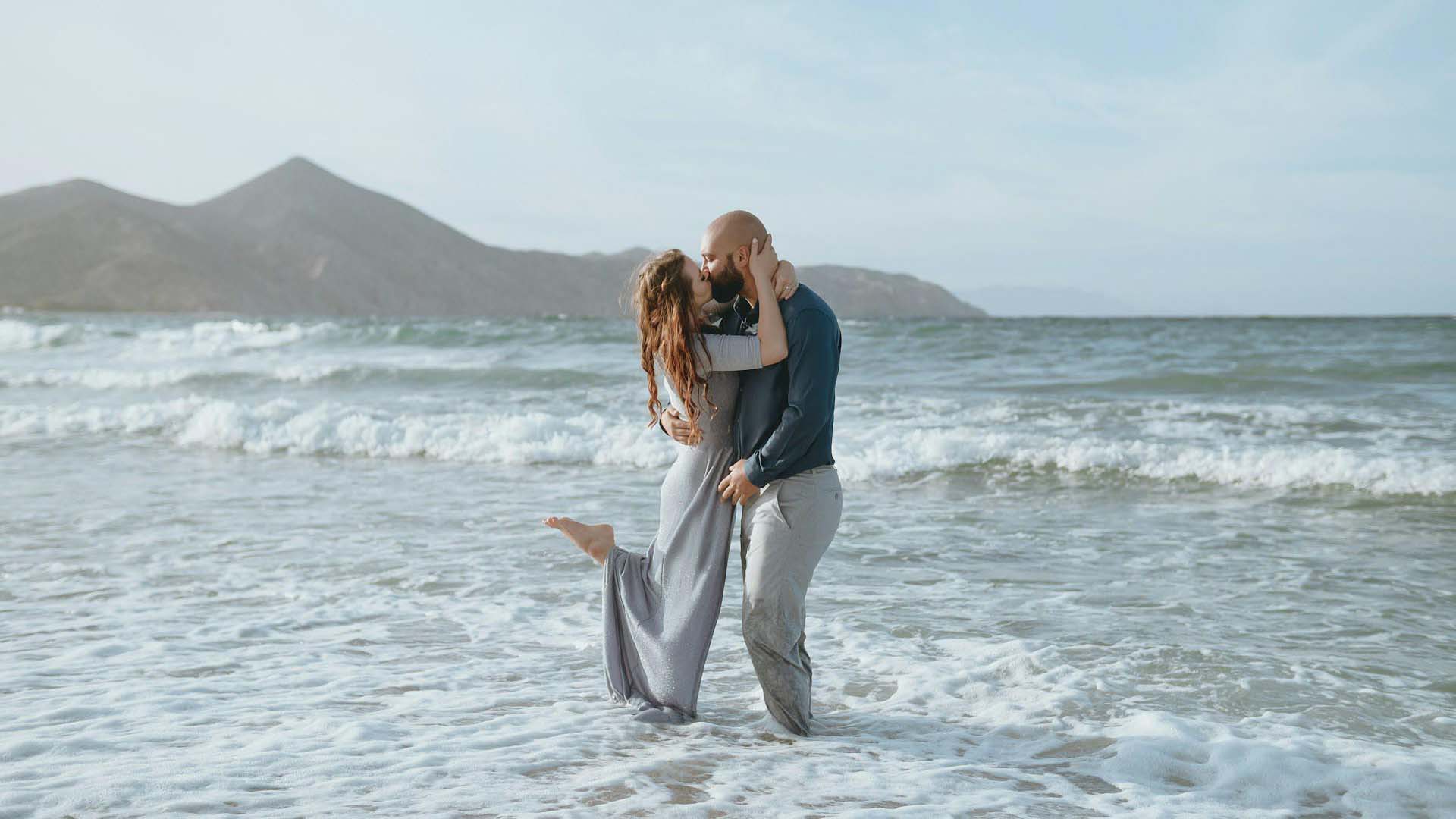 A couple embraces in shallow ocean waters, with gentle waves lapping at their feet and mountains in the background under a blue sky.