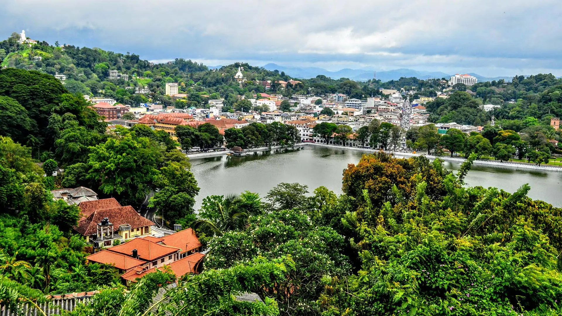 Panoramic view of Kandy City with serene lake, lush greenery, red-roofed buildings, and distant hills under a cloudy sky.