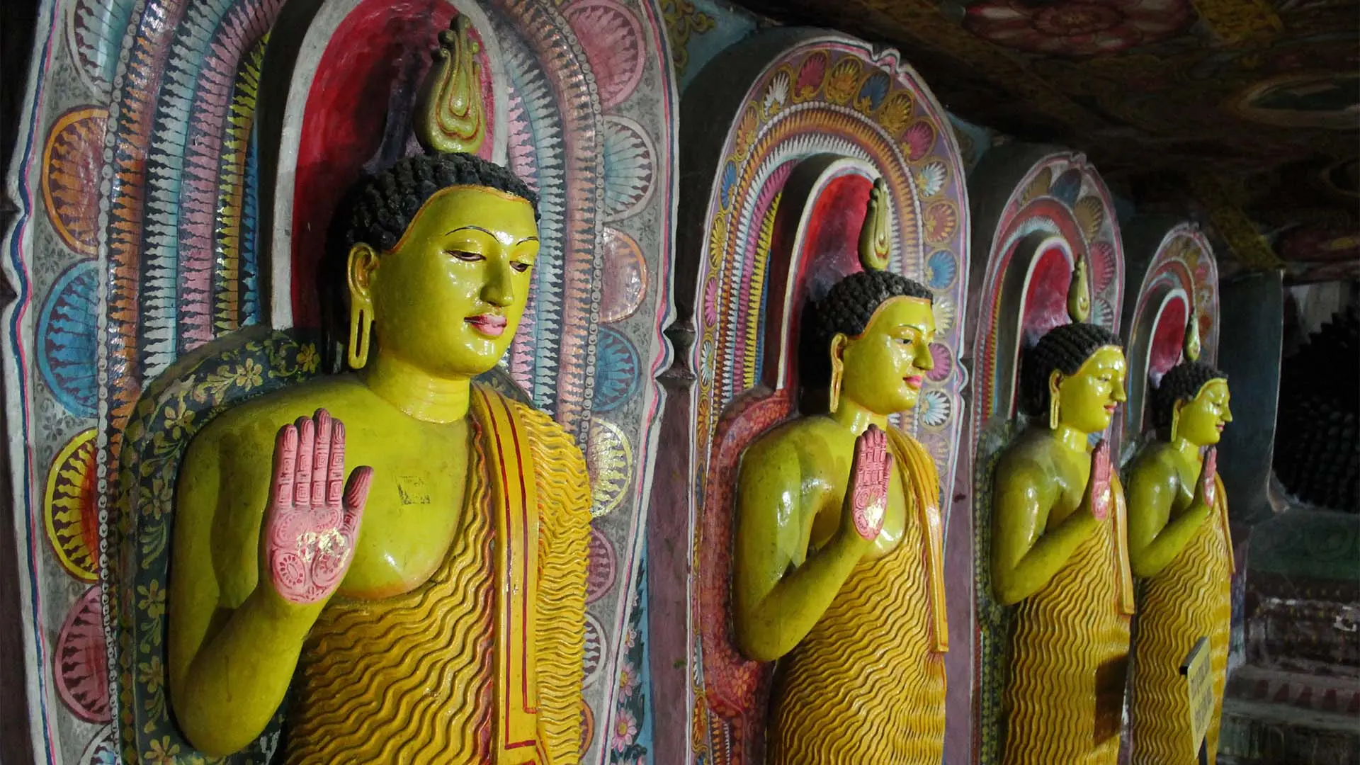 Row of Buddha statues with raised hands, set against colorful backdrops at Aluvihare.