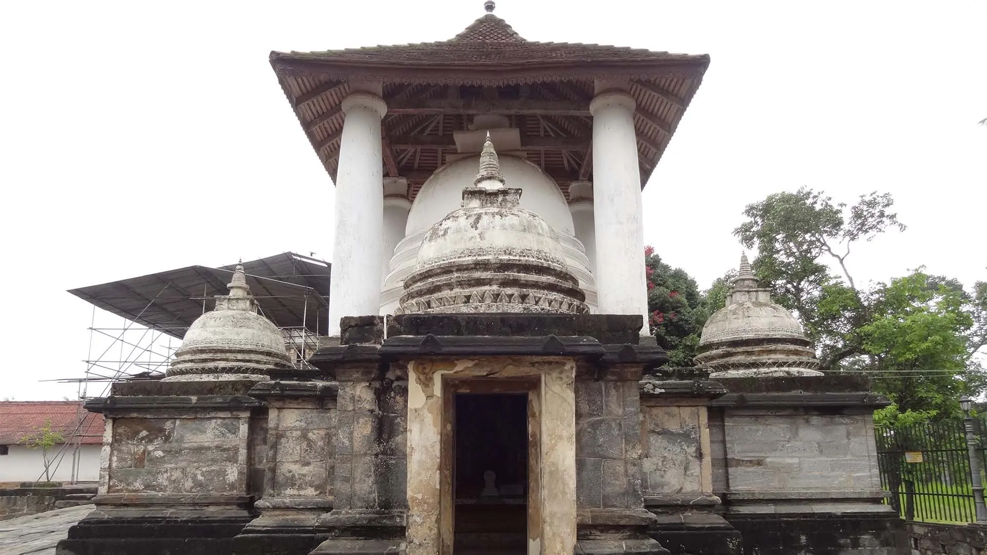 Gadaladeniya Raja maha viharaya with ornate domes and columns, surrounded by lush greenery and a cloudy sky.