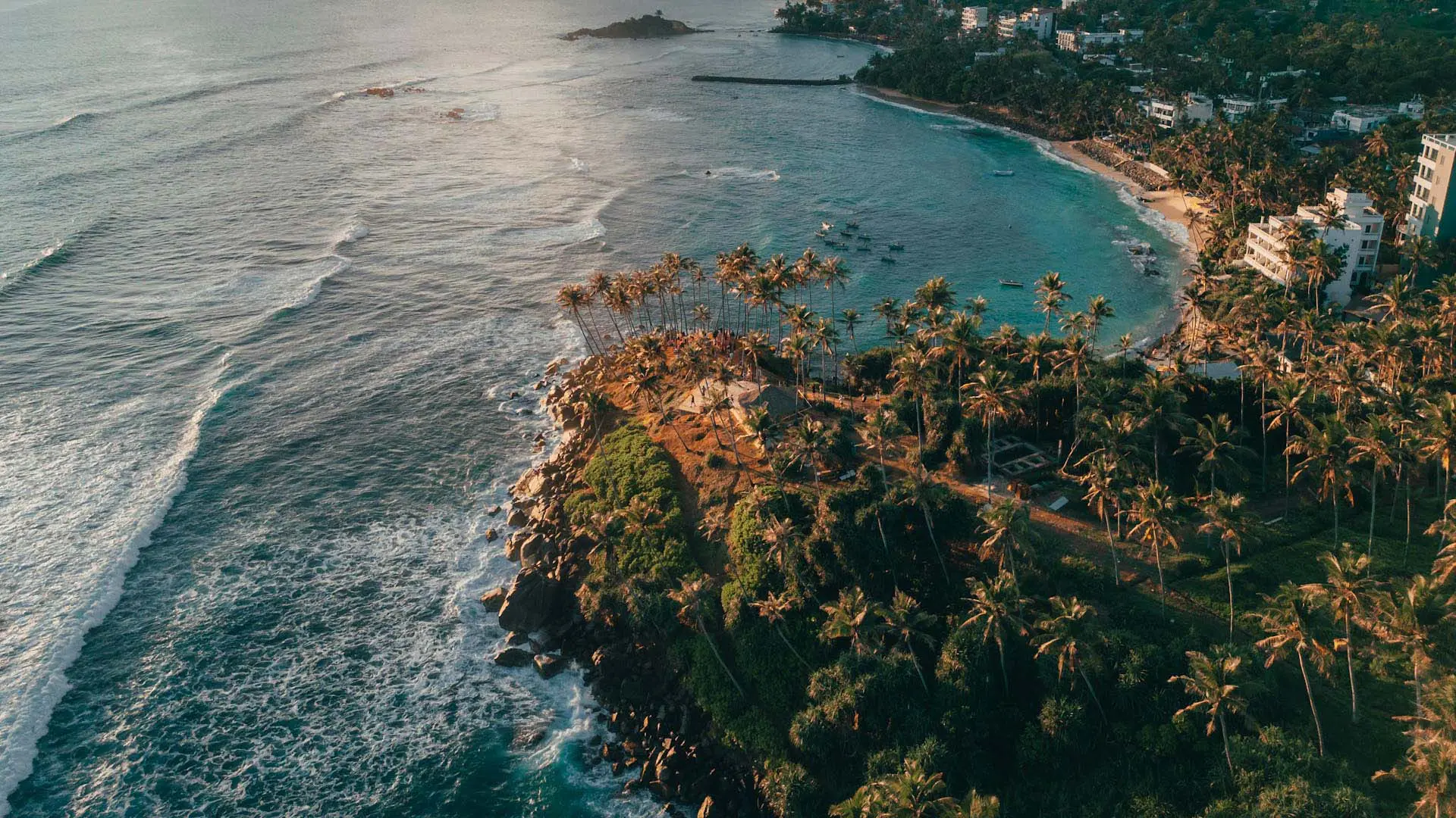 Aerial view of Mirissa Beach with turquoise waters, sandy shores, and a small bay under a partly cloudy sunset sky.