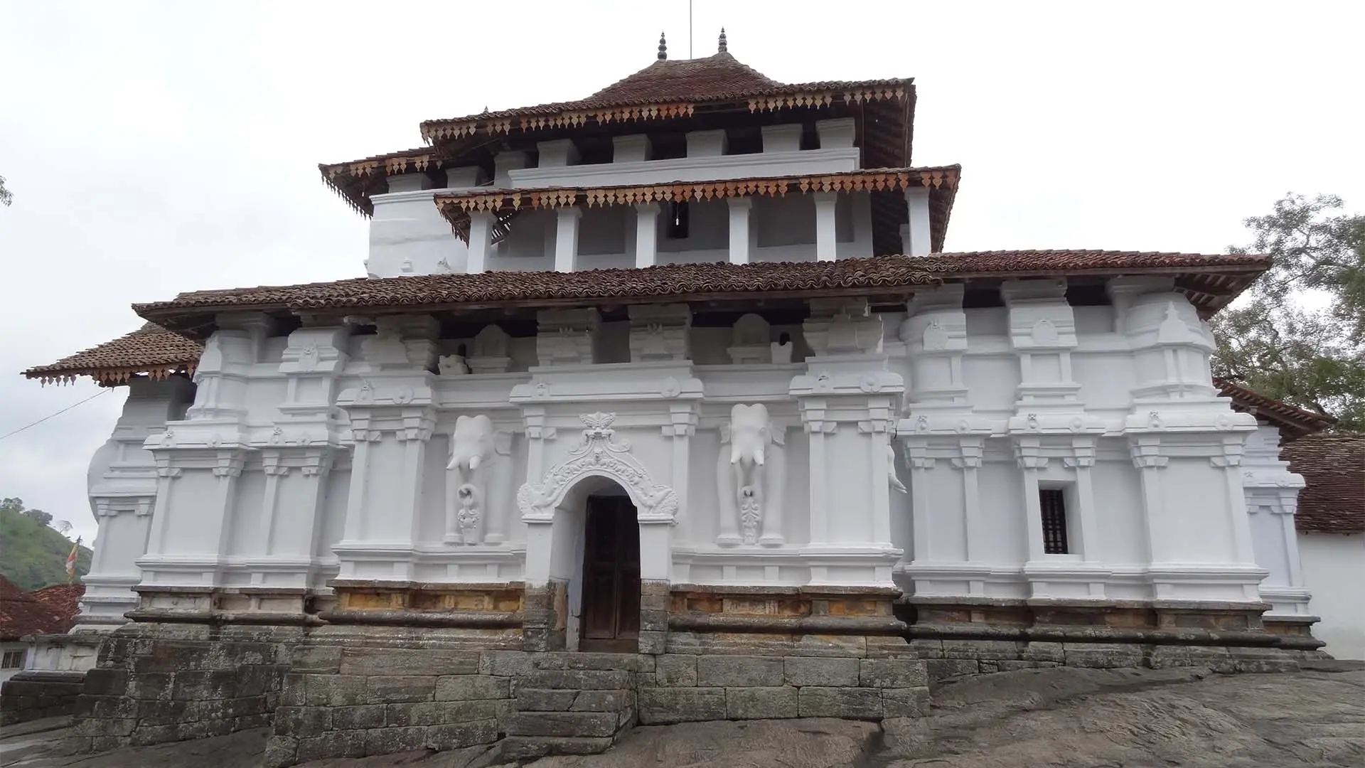Sri Lankathilake Rajamaha Viharaya temple with intricate architecture and red-tiled roof amidst trees under a cloudy sky.