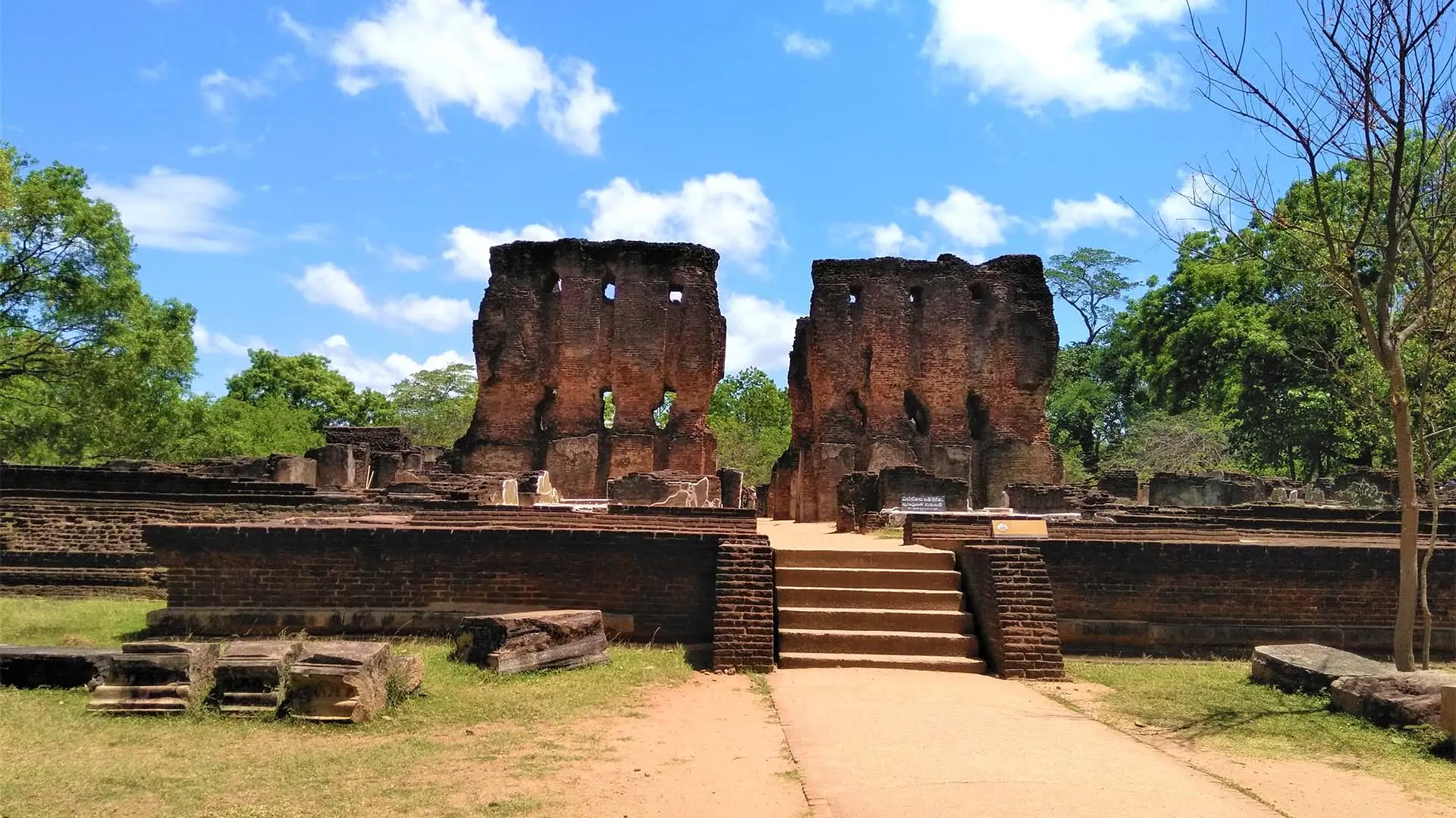 Ancient ruins of the Palace of King Parakramabahu with tall brick walls and a central staircase amidst lush greenery under a clear blue sky.