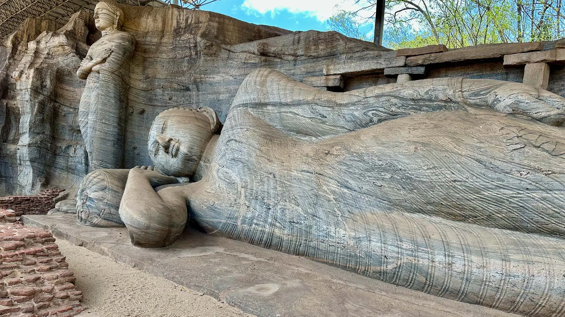 Ancient rock-carved Buddha statues of Gal Viharaya, including a reclining Buddha, set against a stone backdrop under a partly shaded canopy.