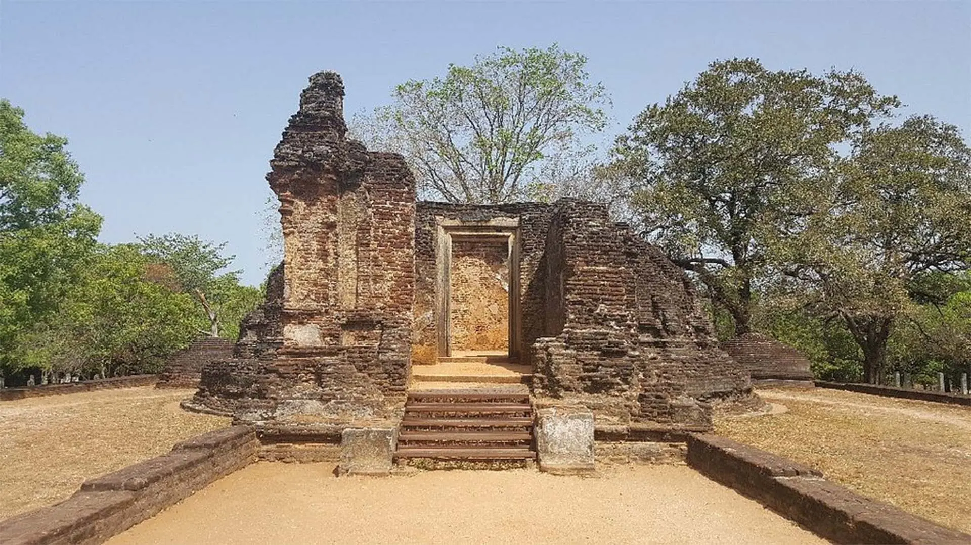 Ancient ruins of Pothgul Viharaya in Polonnaruwa, featuring weathered brick walls and a central entrance, surrounded by lush trees under a clear sky.