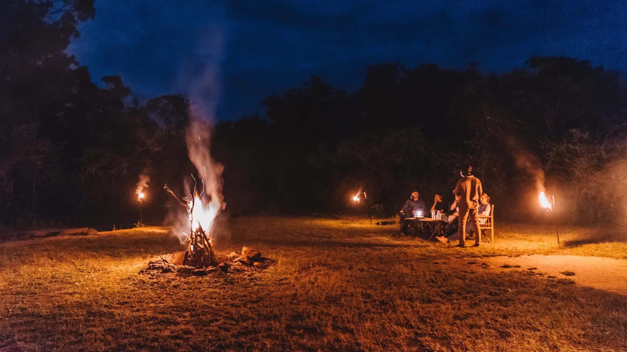 Yala Camping Site at night with a bonfire, people gathered around a table lit by torches, and surrounding forest under a dark sky.